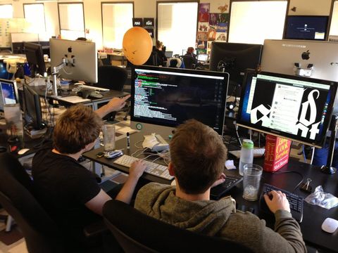 Zwei Entwickler von hinten vor Monitoren, einer zeigt auf Code im Terminal